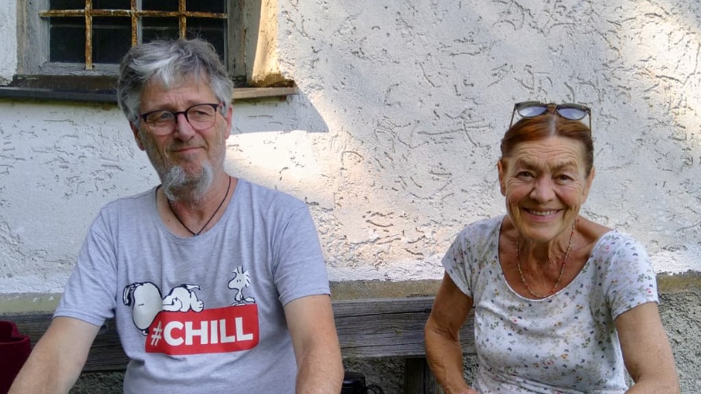 Zwei Menschen sitzen auf einer Holzbank vor einer weißen Putzwand, freundlich in die Kamera blickend – Bettina und Jochen Nistler, die kesslfligga.