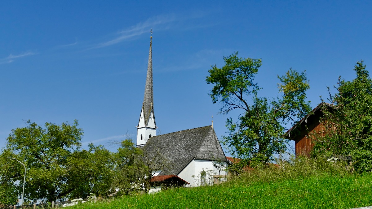 Kirche St. Jakobus in Bernhaupten mit spitzem Turm, umgeben von Bäumen und Wiese unter blauem Himmel.