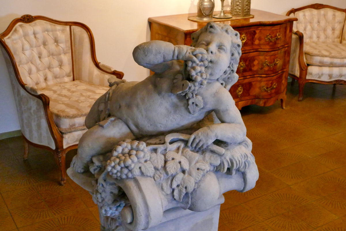 Barocke Puttenstatue, die entspannt auf einem Sockel liegt und eine Traube zum Mund führt; im Hintergrund zwei gepolsterte Sessel und eine Kommode im Barockstil.